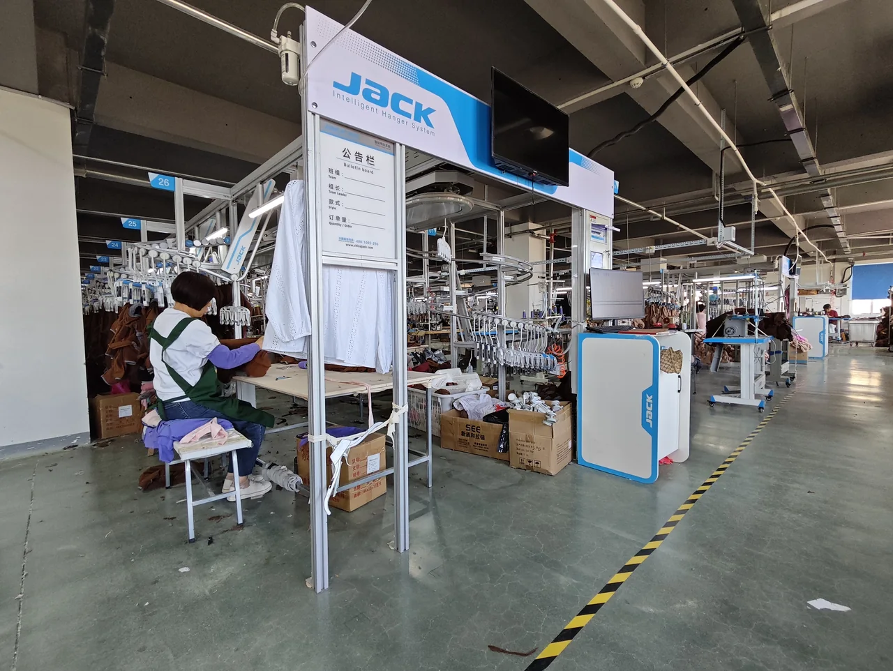 Worker at a sewing station beside an overhead garment handling line and marked aisle